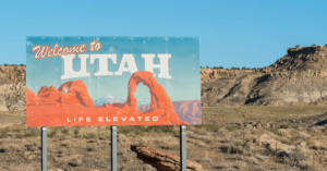 A "Welcome to Utah" sign featuring Delicate Arch and the slogan "Life Elevated" stands against a backdrop of arid desert landscape and blue sky.