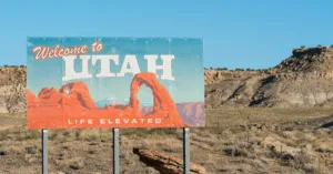 A "Welcome to Utah" sign featuring Delicate Arch and the slogan "Life Elevated" stands against a backdrop of arid desert landscape and blue sky.